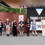 A group of people stand on a stage with a sign behind them that says "Trans Pride"